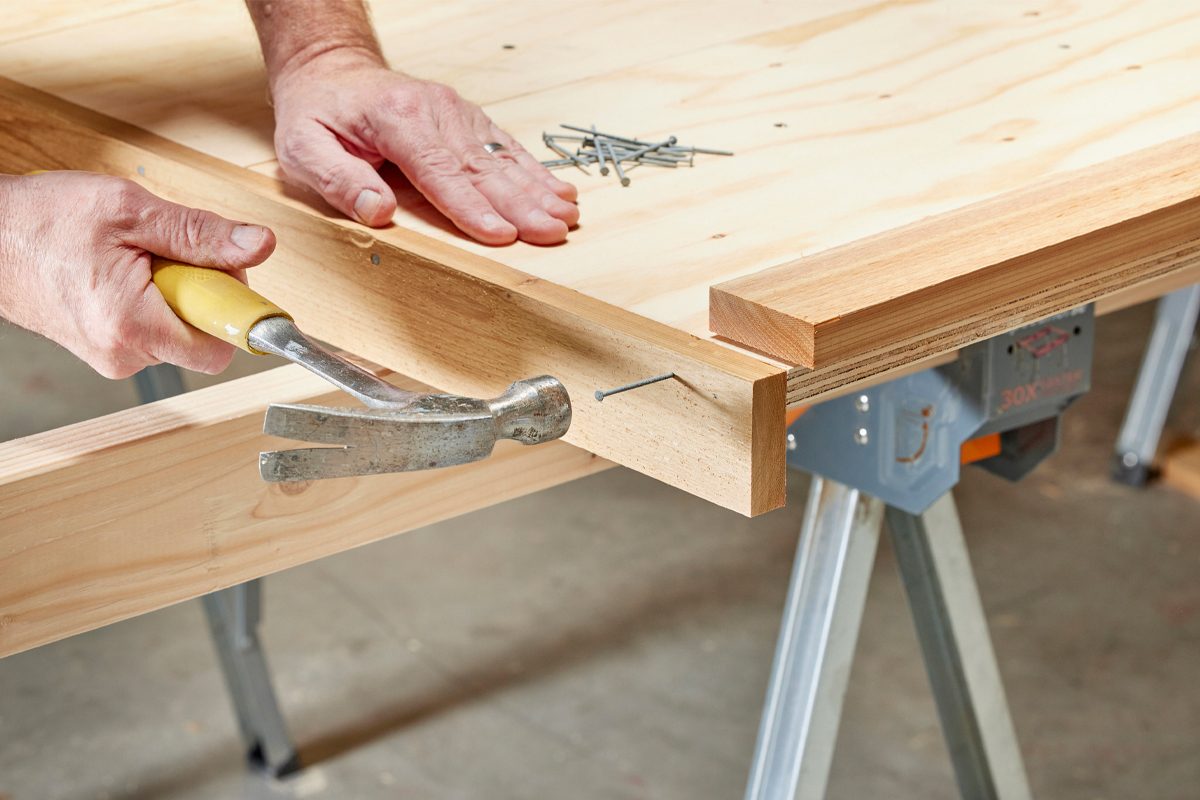 A hand with a yellow-handled hammer drives a nail into wooden planks, with scattered nails nearby, on a workbench in a workshop setting.