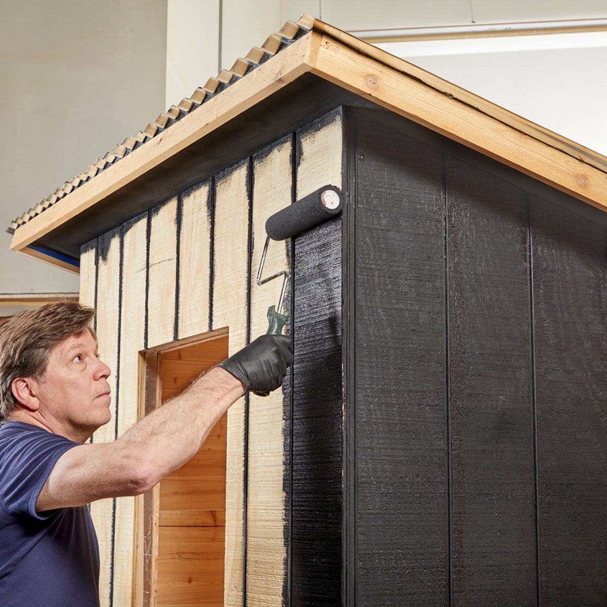 A man uses a paint roller to apply black paint on the side of a wooden structure, under a sloped roof, in a well-lit indoor environment.