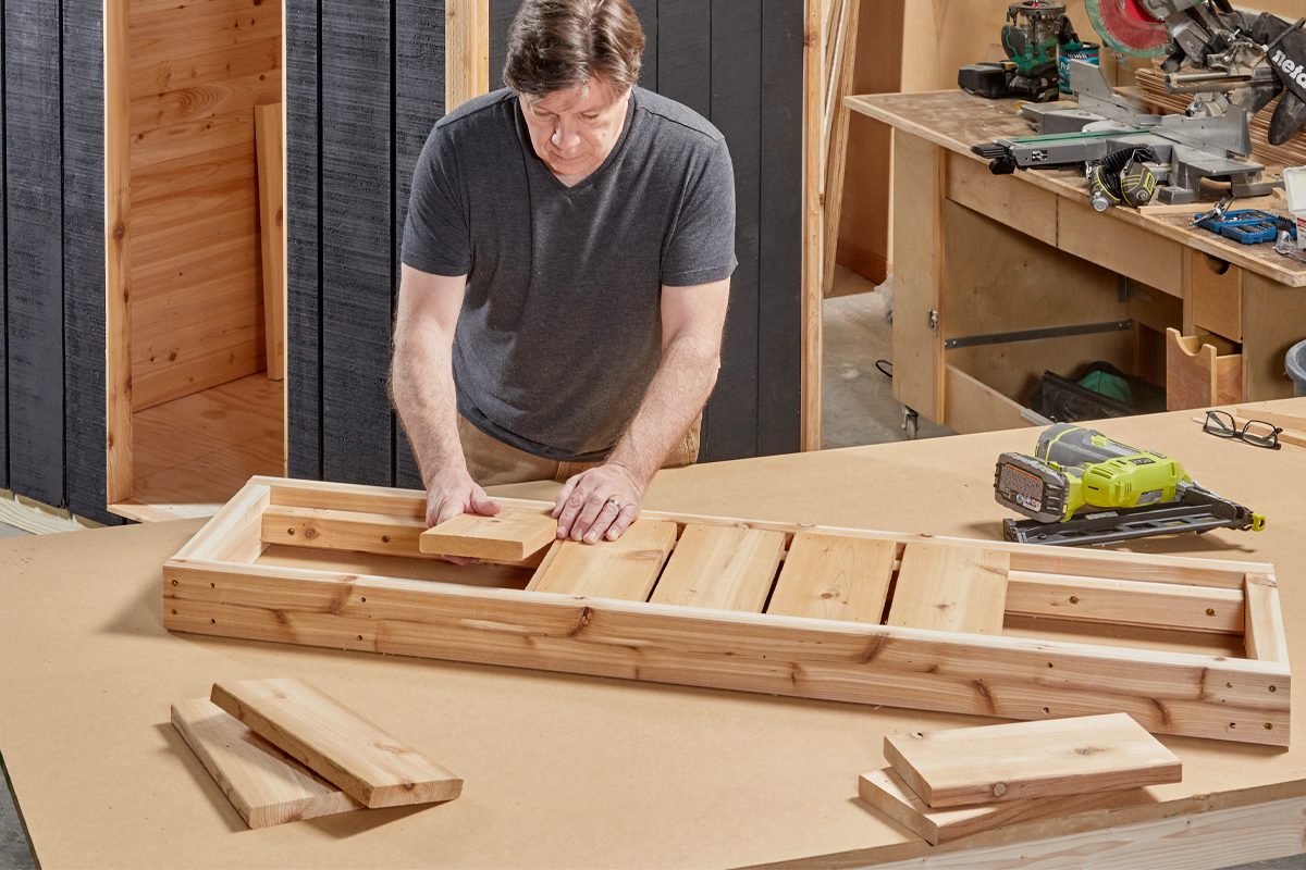 A man places wooden planks into a rectangular frame on a workbench in a workshop filled with tools and wooden materials.