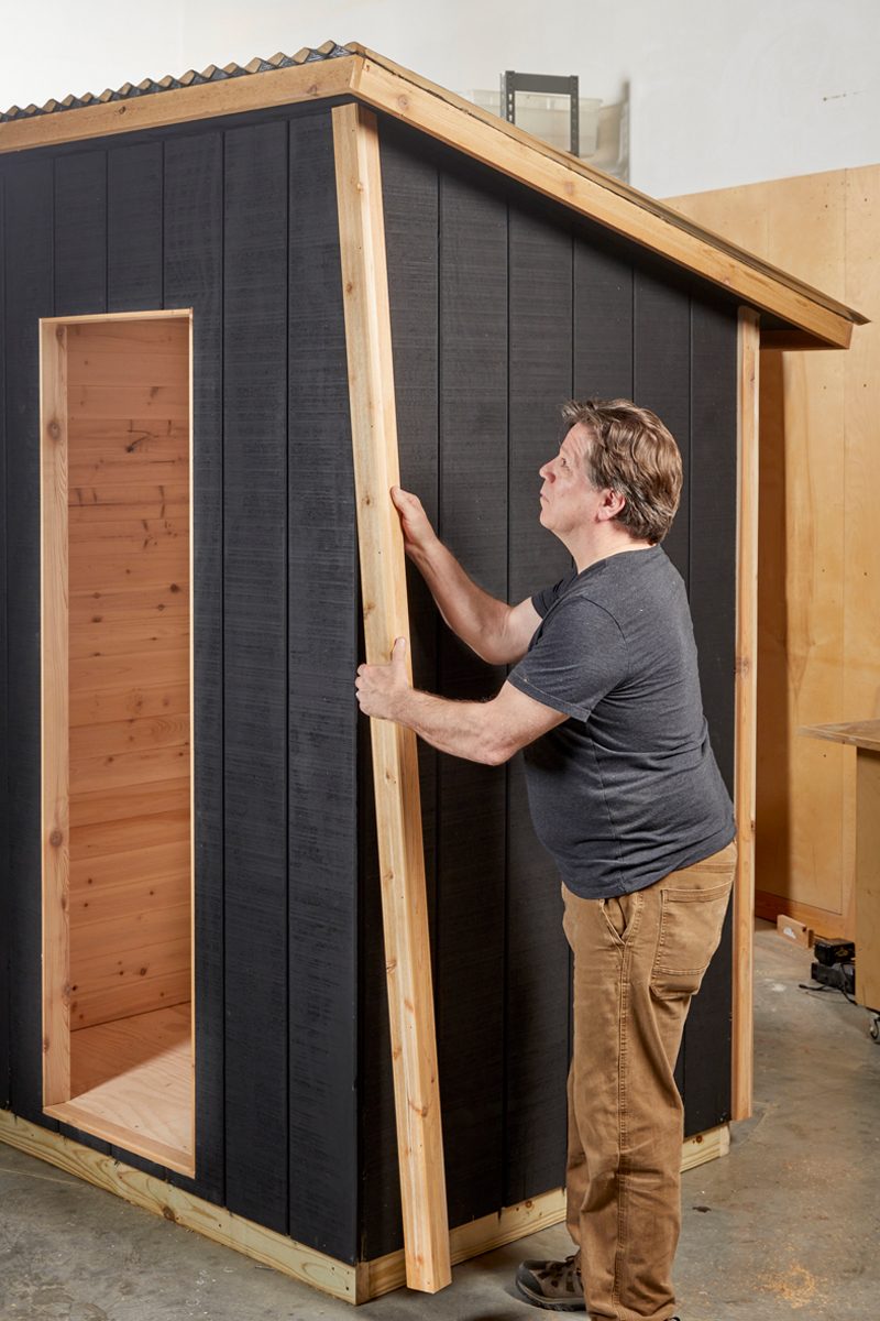 A man is securing a wooden frame against the black exterior of a partially constructed shed in a workshop environment.