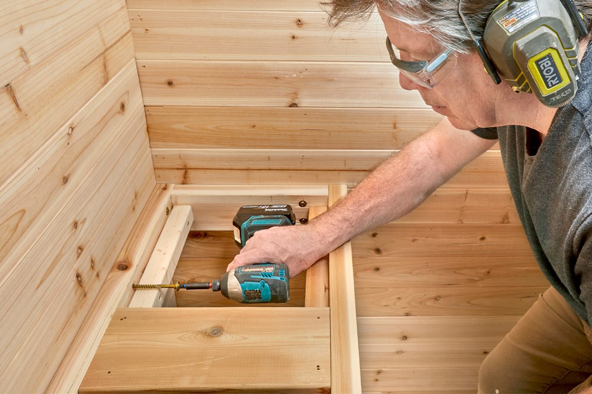 A person drives a screw into wooden planks using a power drill, surrounded by wooden walls in a workshop or construction space.