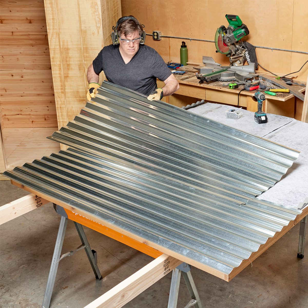 A man in protective gear lifts a sheet of corrugated metal in a workshop, surrounded by tools and wooden surfaces, preparing for a project.