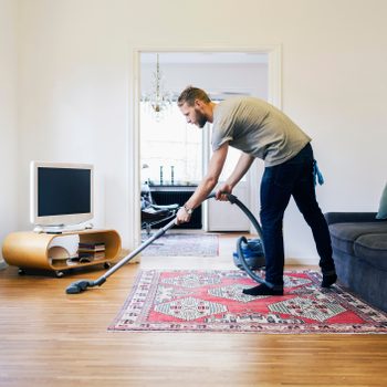 A man vacuums a patterned rug in a bright living room, surrounded by a modern television stand and a couch, with a soft glow from nearby windows.