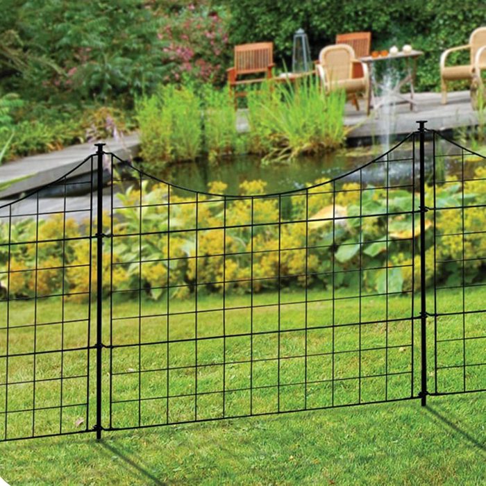 A black grid fence stands on green grass, separating a tranquil pond with lush plants and garden furniture in the background.