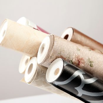 Rolls of decorative wallpaper lean against each other, showcasing various patterns and colors, in a minimalistic, well-lit environment.