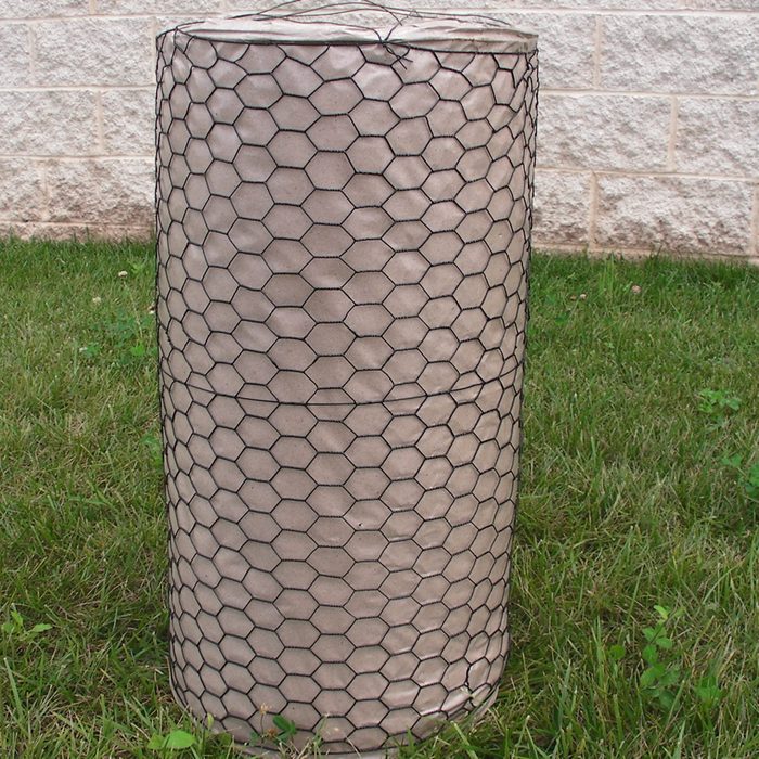 A cylindrical object, covered in hexagonal wire mesh, stands on grass beside a textured stone wall. It appears to be a garden or construction material.