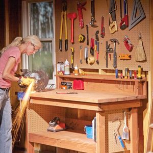 A person operates a power saw, generating sparks, at a wooden workbench surrounded by various tools mounted on a pegboard in a workshop.