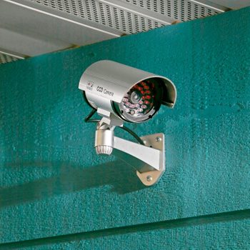 A security camera is mounted on a teal wall, positioned to monitor the area. It has a cylindrical shape with a silver finish and visible red lights.
