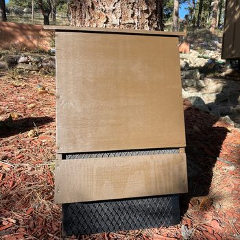 A wooden structure stands against a tree, partially open at the bottom, surrounded by red mulch and natural scenery, with shadows cast by nearby trees.