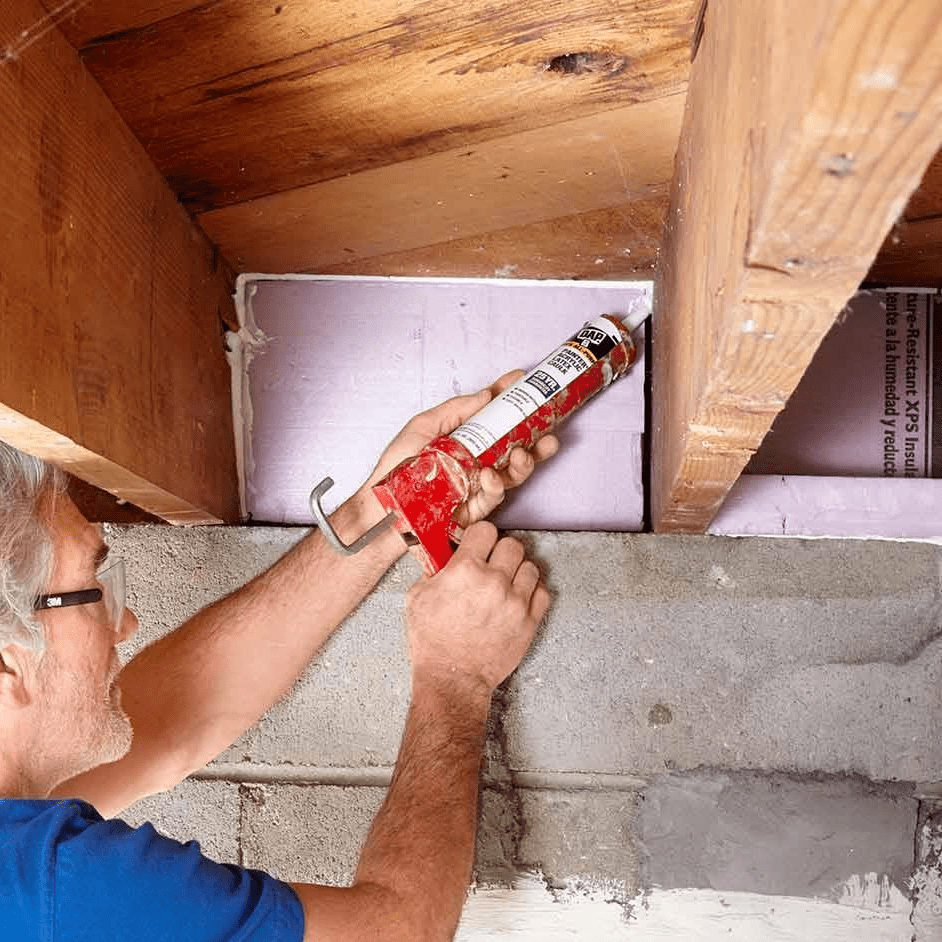 A person uses a caulking gun to apply sealant in a construction area, surrounded by wooden beams and insulated wall panels.