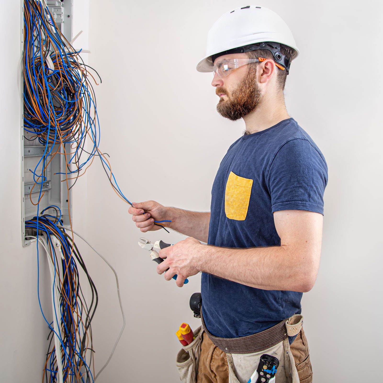A man in a hard hat and safety glasses works with electrical wires using pliers, while surrounded by a tangle of colorful cables in a neutral-colored room.