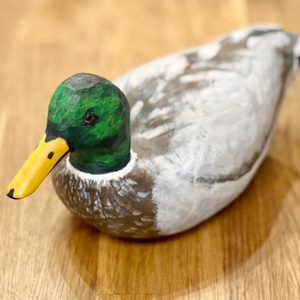 A painted wooden duck sits on a wooden surface, showcasing vibrant green and yellow colors, resembling a mallard. It appears stationary in a cozy indoor setting.