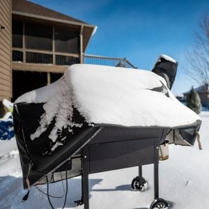 A grill covered in snow sits on a snowy yard, with a clear blue sky above and a house with a screened porch in the background.