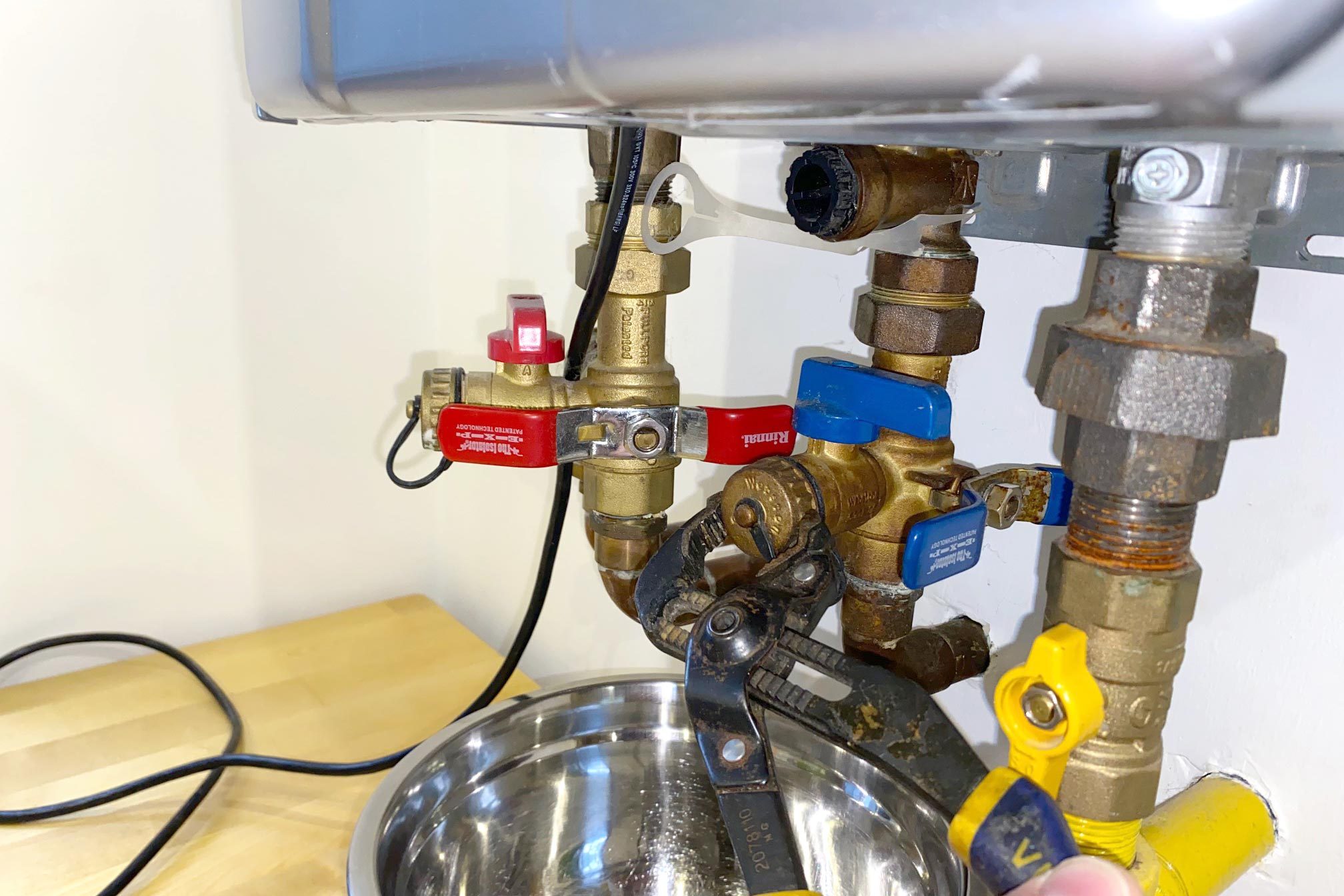 A person uses pliers to manipulate plumbing valves connected to a metal bowl on a wooden surface in a well-lit kitchen environment.