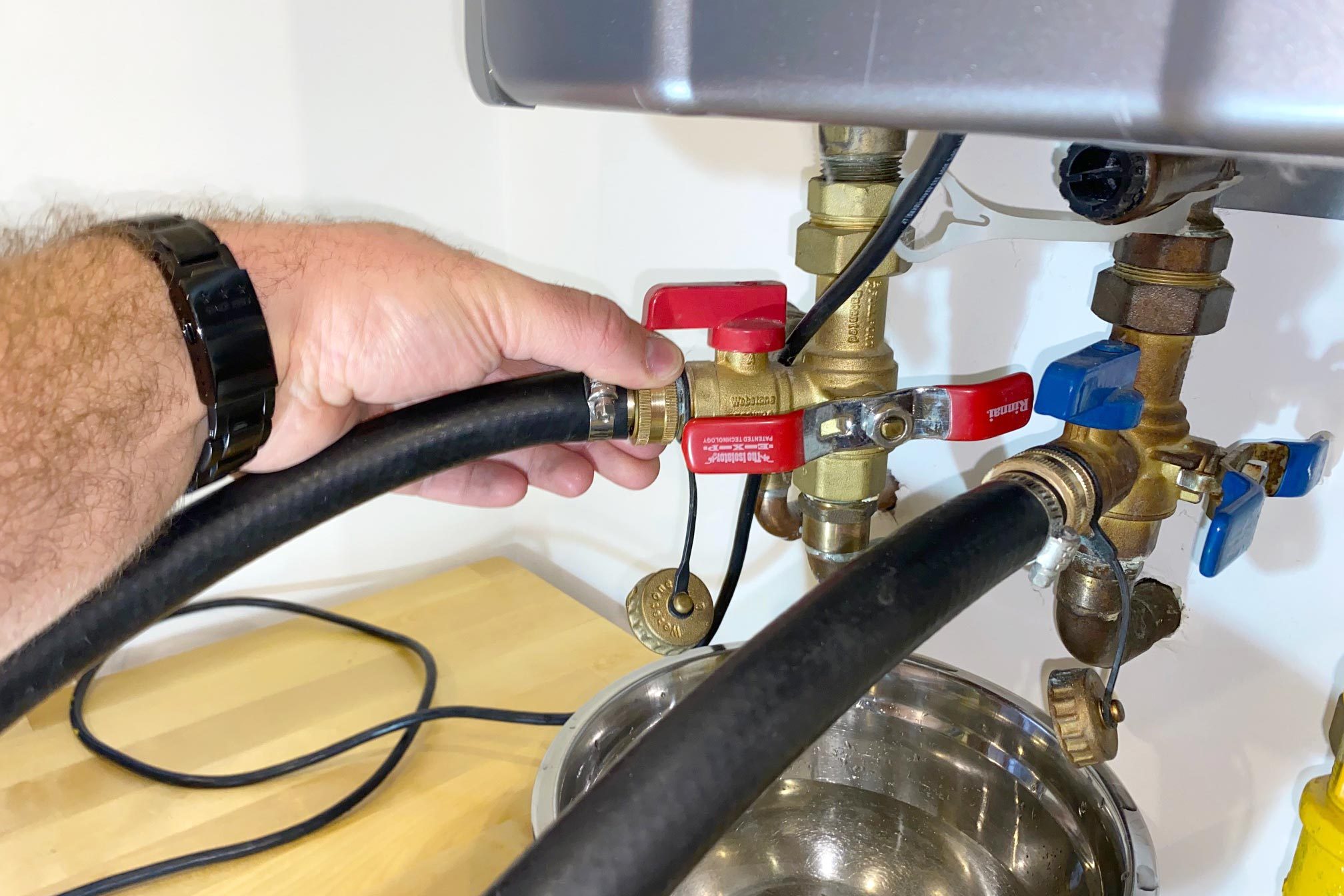 A hand grips a black hose connected to a brass valve, adjusting it near a metal bowl on a wooden surface in a domestic setting.