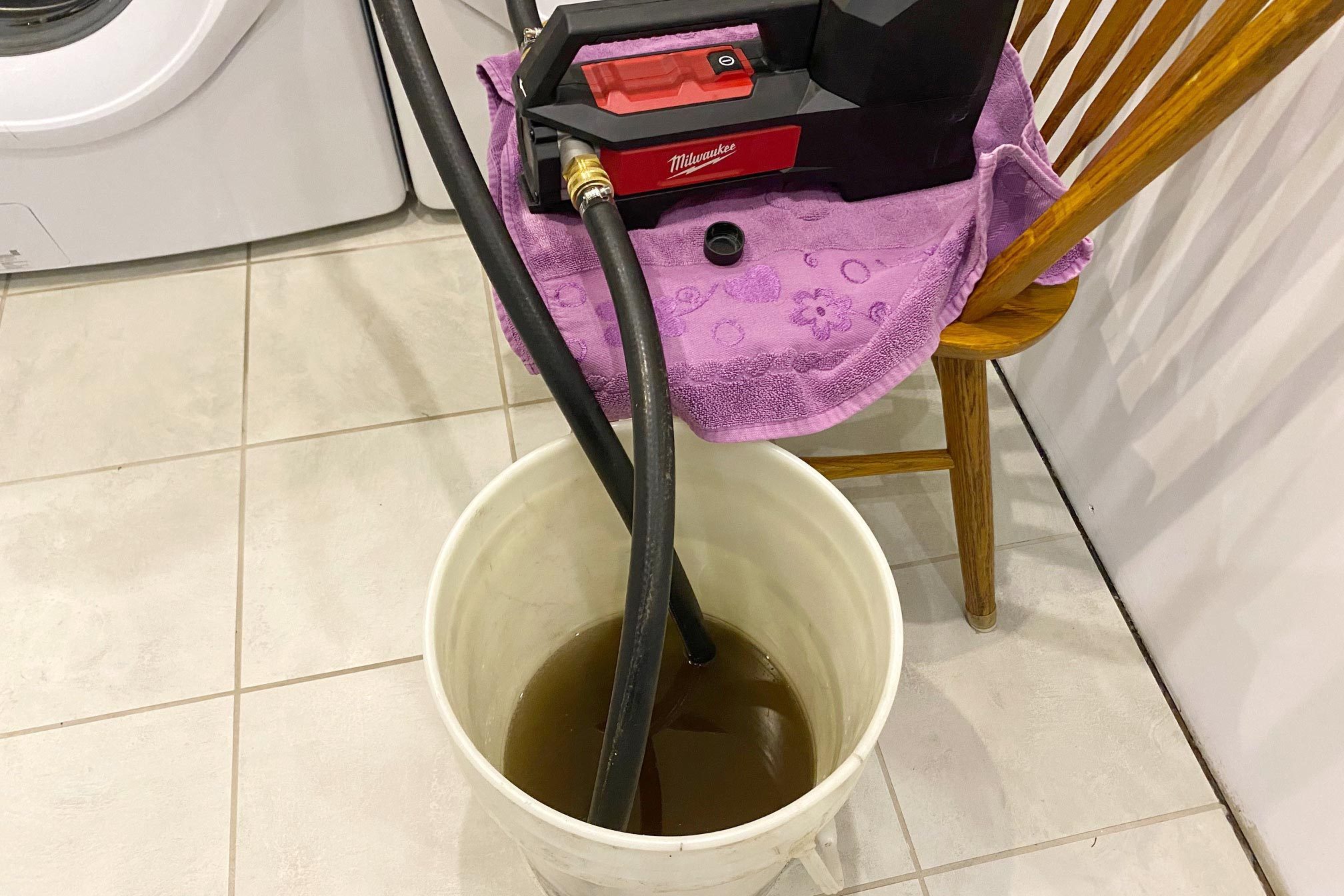 A pump sits on a towel-covered chair, connected to hoses that drain murky liquid into a white bucket on a tiled floor.