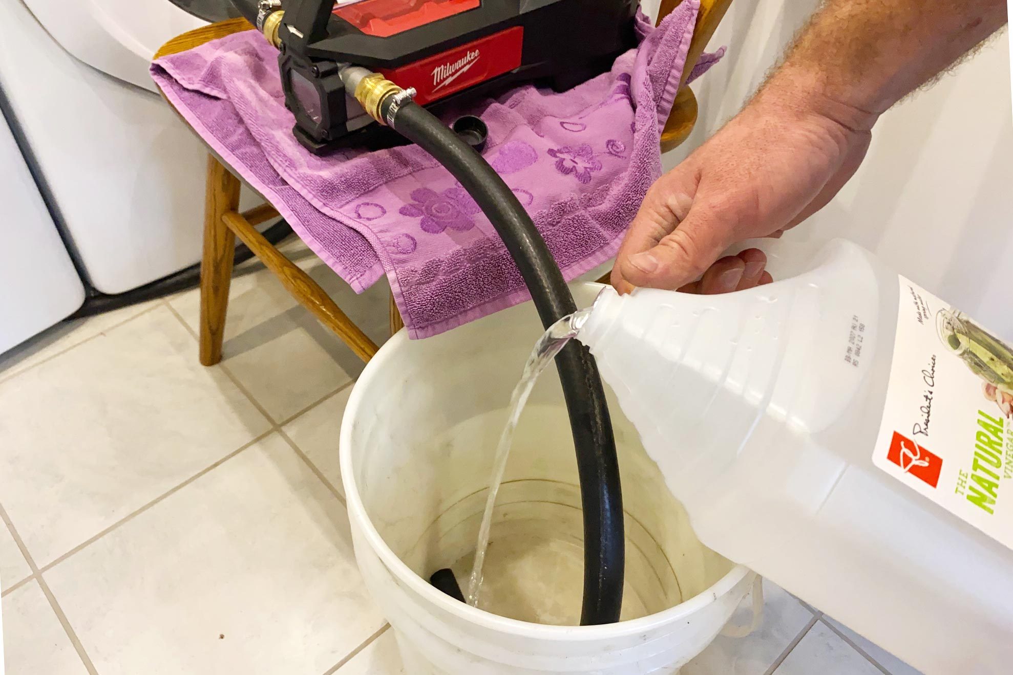 A person pours liquid from a bottle into a bucket through a black hose, set on a towel-covered chair in a tiled laundry room.