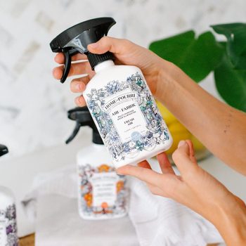 A hand holds a white spray bottle labeled "Home-Pourri Air + Fabric." It's surrounded by other similar bottles on a light surface, with green leaves in the background.