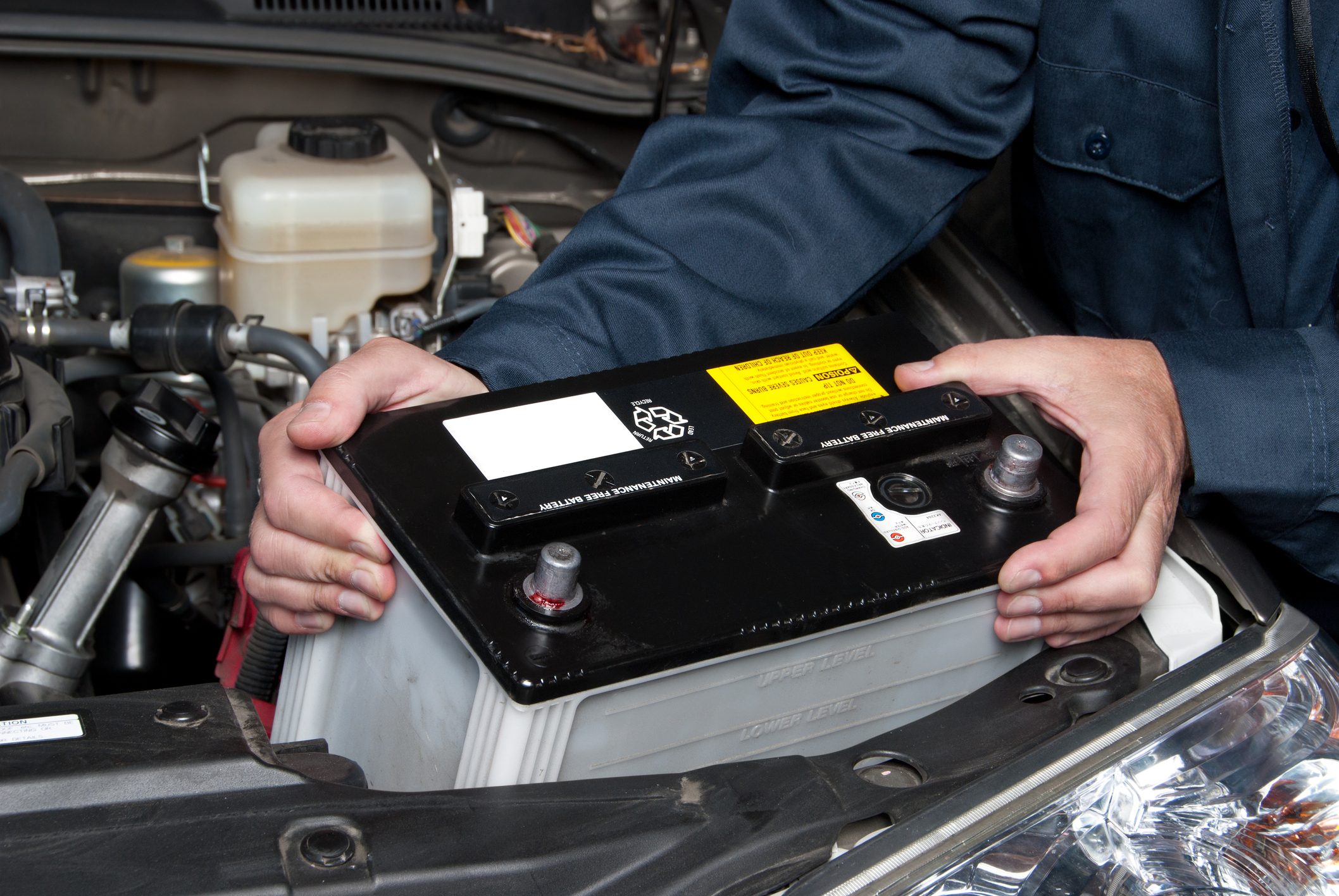A person is installing a car battery under the hood, surrounded by engine components and a nearby reservoir, focusing on proper alignment and secure placement.