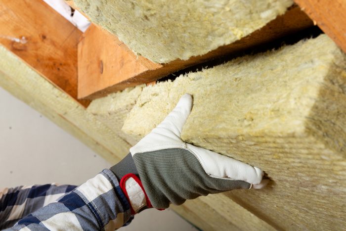 A gloved hand presses insulation against wooden beams in a construction setting, configuring material for installation in a wall or ceiling.