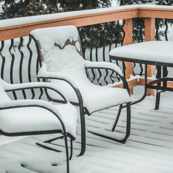 Two snow-covered metal chairs and a table sit on a wooden deck, surrounded by falling snowflakes and a snowy landscape in the background.