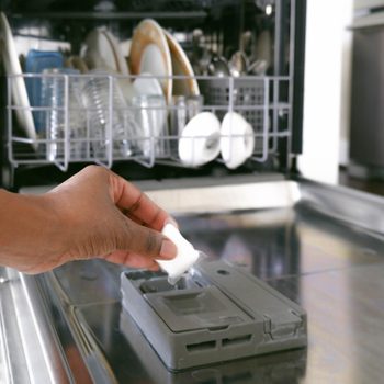 A hand adds a dishwasher pod into the detergent compartment of an open dishwasher, which contains clean dishes and glassware.