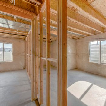 Framed walls stand in an unfinished interior space, with sunlight streaming through windows and a concrete floor, suggesting ongoing construction.