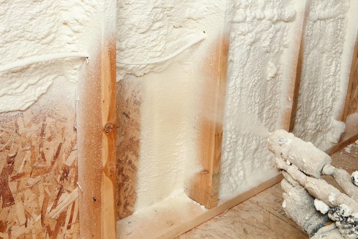 A spray foam insulation gun is applying a thick layer of foam onto wooden wall studs in a construction environment.