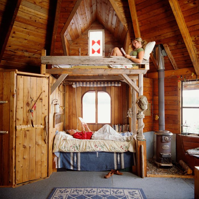 A wooden loft bed holds a woman reading, while a man rests below. The cozy cabin features a window, a stove, and rustic decor.