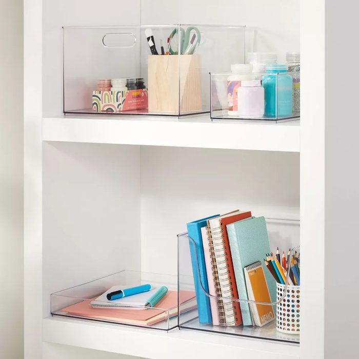 Clear storage bins hold various craft supplies and stationery on white shelves, showcasing organized items like books, notebooks, and art materials in a tidy setting.
