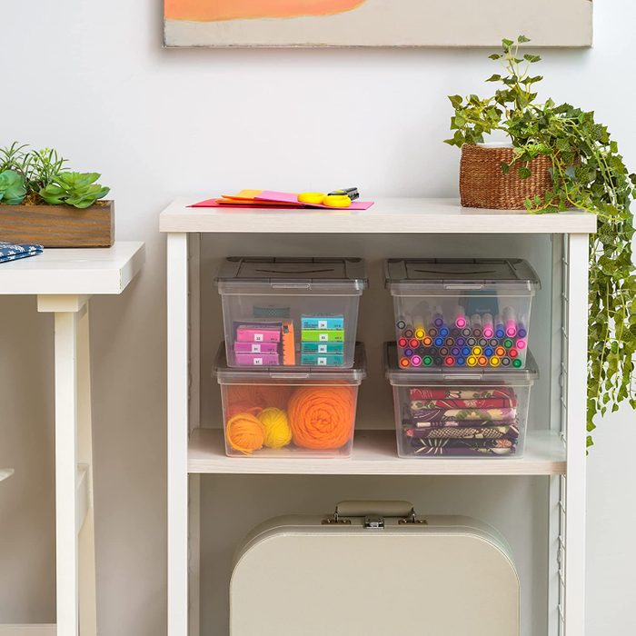 Clear storage containers hold various craft supplies on a shelf, while a decorative plant and stationery are arranged on the table above in a bright room.