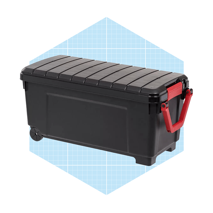 A black storage container with a red handle rests on a light blue grid background, showcasing its robust design and wheels for easy movement.