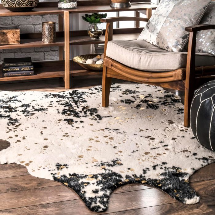 A patterned cowhide rug lies on wooden flooring, under a wooden chair with a cushion. Nearby, a shelf displays decorative items and books.