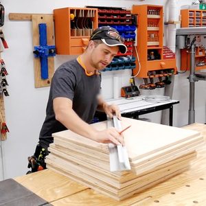 A man is measuring a stack of plywood sheets on a workbench in a well-equipped workshop filled with tools and materials.
