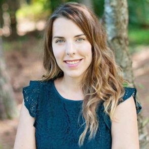 A woman with long, wavy hair smiles while seated outdoors near trees, wearing a dark dress with decorative patterns. Natural, soft lighting enhances the scene.