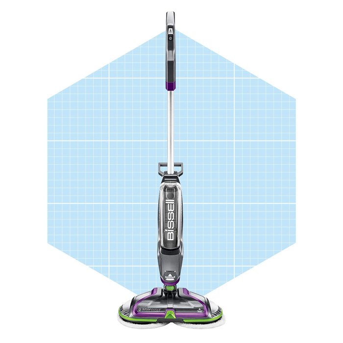 A floor cleaner stands upright with a sleek design, actively ready to clean surfaces, set against a light blue geometric background.