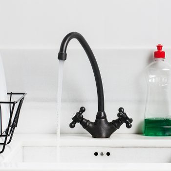 A black faucet streams water into a white sink, beside a green dish soap bottle and a wire dish rack holding plates in a bright kitchen.