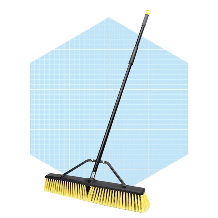 A large broom stands upright, featuring a long handle and wide bristles, set against a light blue grid background.
