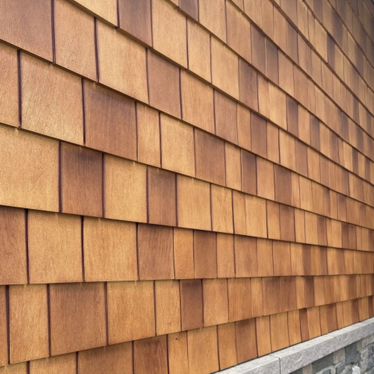 Wooden shingles cover a wall, arranged in overlapping rows. A stone foundation borders the base. The surface appears smooth with a warm, golden hue.