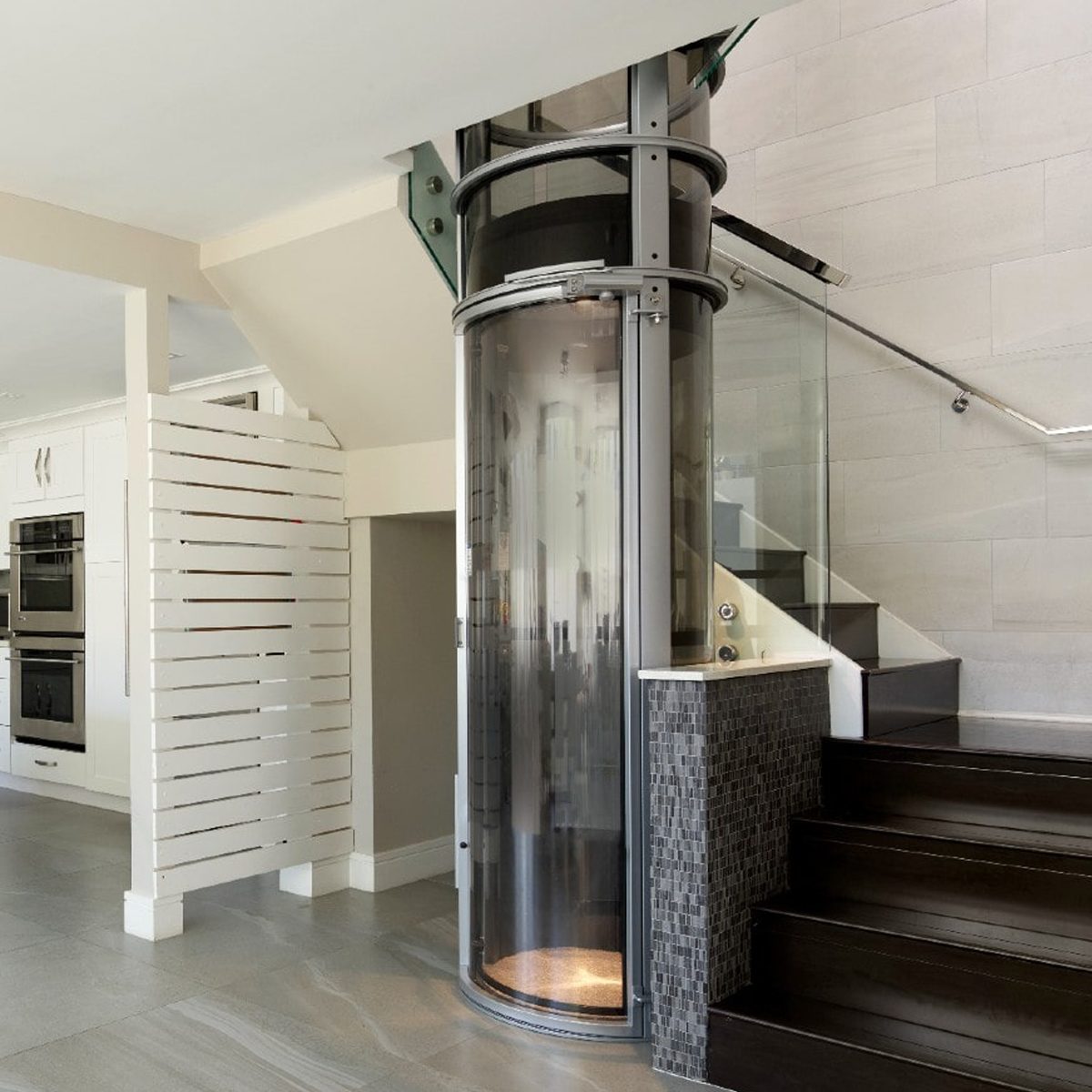 A circular glass elevator is stationed beside dark wooden stairs, connecting different levels in a modern interior space with sleek, minimalist design elements.