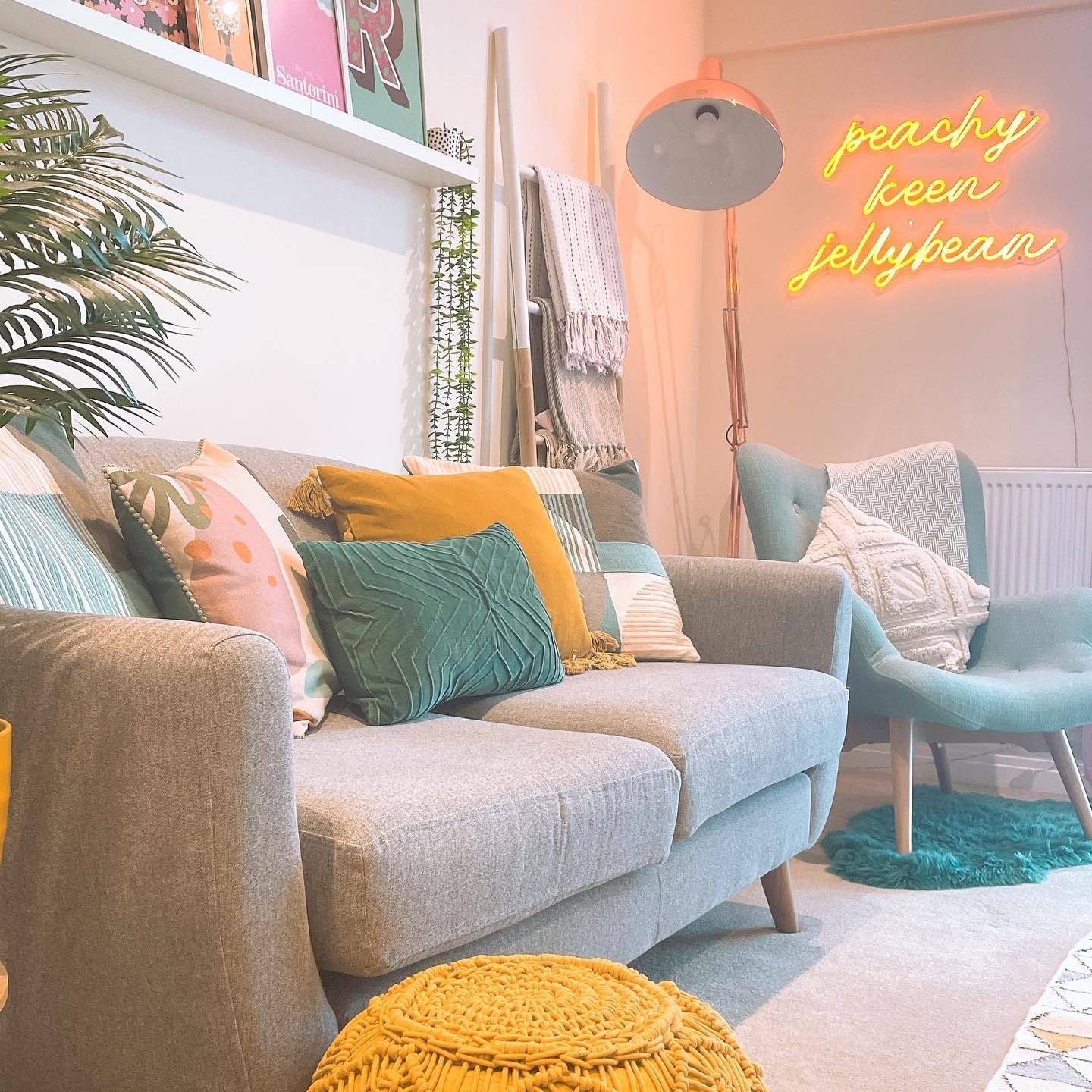 A cozy living room features a gray sofa with colorful pillows, a turquoise chair, a yellow pouf, and neon wall art in a bright space.
