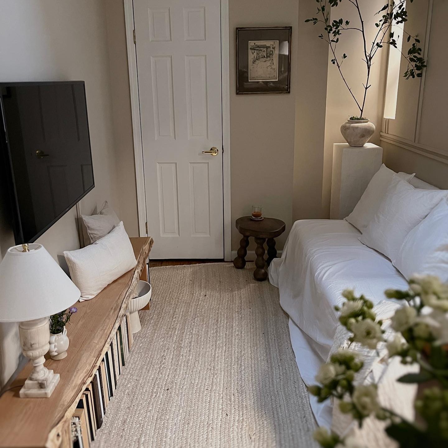 A white sofa sits in a cozy room with a wooden bench, a plant, and a TV on the wall, creating a serene living space.