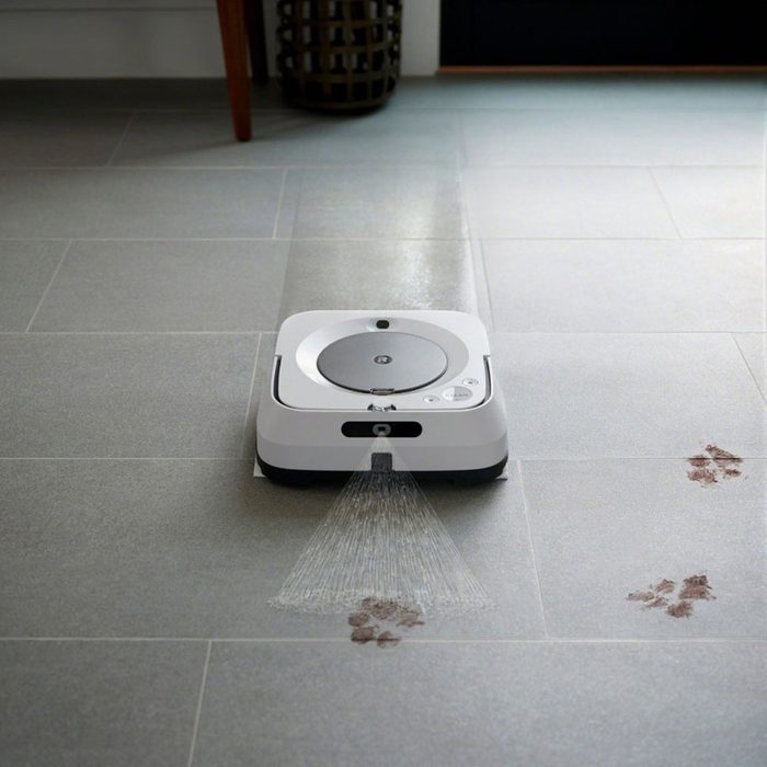 A robotic cleaner sprays water as it moves across gray tile flooring, targeting brown paw prints nearby, with a basket and door in the background.
