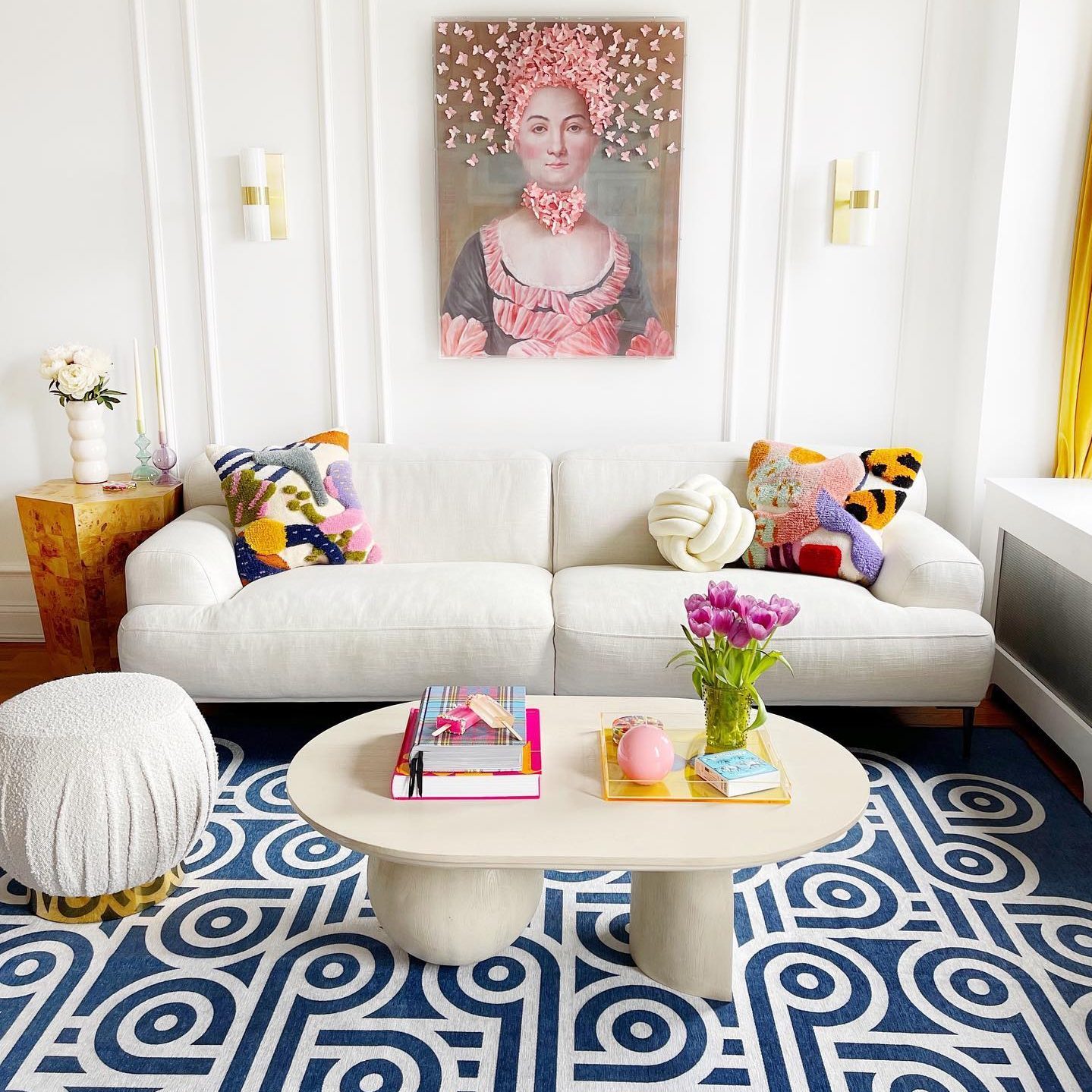 A white sofa sits in a brightly lit room, adorned with colorful pillows. A round coffee table displays books and flowers, with patterned rugs beneath.