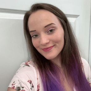 A person with long, dark hair featuring purple highlights smiles at the camera, wearing a floral-patterned shirt, against a plain background.