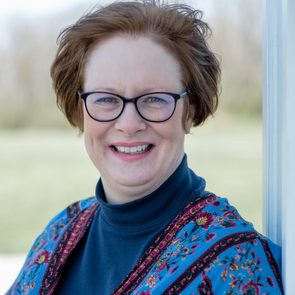 A woman with short, wavy hair smiles while wearing glasses and a colorful patterned shawl, standing outdoors against a blurred green background.
