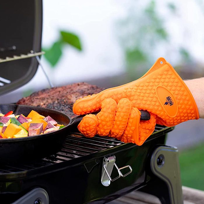 An orange silicone glove grips a skillet of colorful vegetables above a grill, with a cooked piece of meat nearby in an outdoor setting.