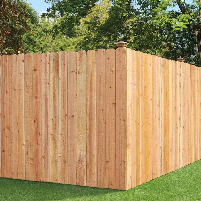 A wooden fence stands tall, enclosing a space, with green grass below and trees in the background under a clear sky.