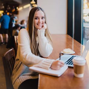 A young woman sits at a cafe table, smiling while writing in a notebook, with a laptop and a coffee cup beside her, and a pastry nearby.