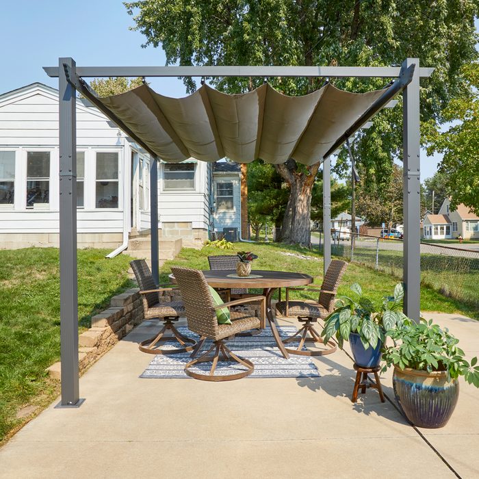 A patio table surrounded by wicker chairs sits under a fabric canopy, near a house and trees, with potted plants decorating the space.